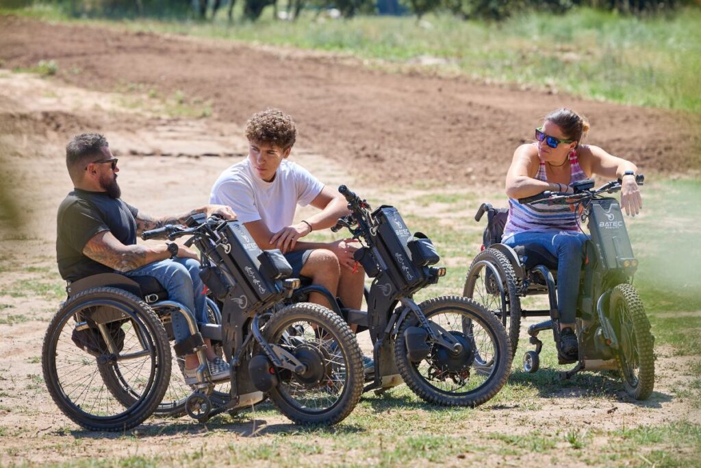 Wheelchair users with electric handbikes on a dirt path.