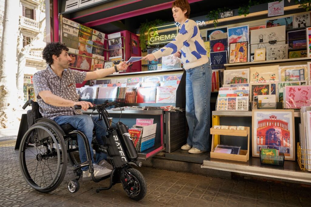 Wheelchair user with an electric handbike at a kiosk.