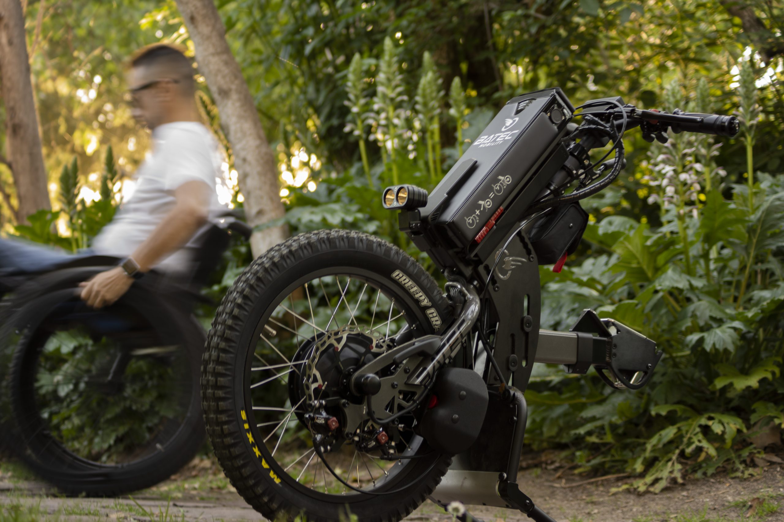 Fotografía del catálogo del handbike eléctrico todoterreno Batec Scrambler 2 desanclado con usuario en silla de ruedas de fondo.