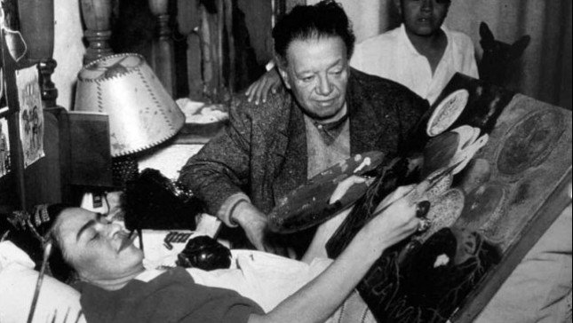 Frida Kahlo tumbada en su cama junto a Diego Rivera pintando.